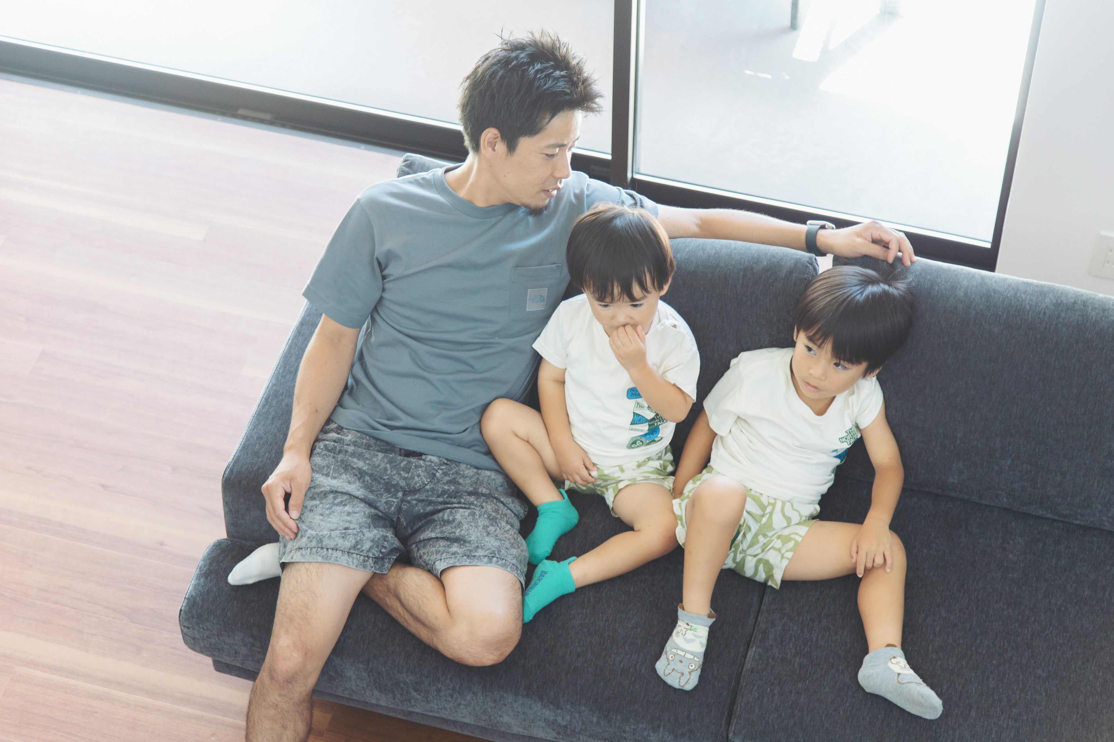 A man and two children sitting on a grey sofa in a room with a large window.