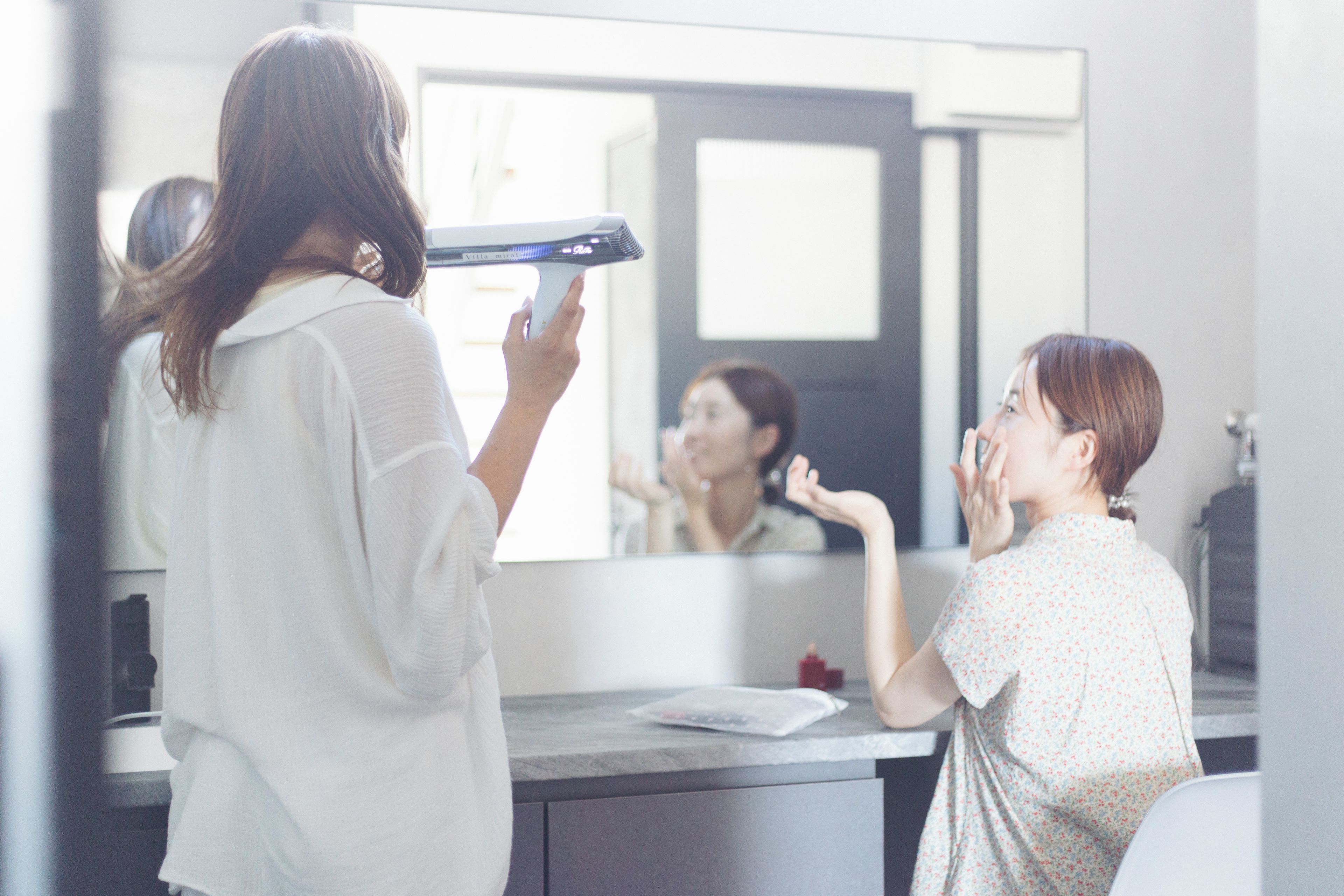 A person using a hairdryer in a bathroom with a mirror, a sink, and a window.