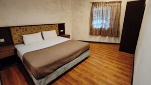 Un dormitorio con piso de madera, una cama con cubierta marrón, una mesita de noche, una ventana con cortinas y una puerta.