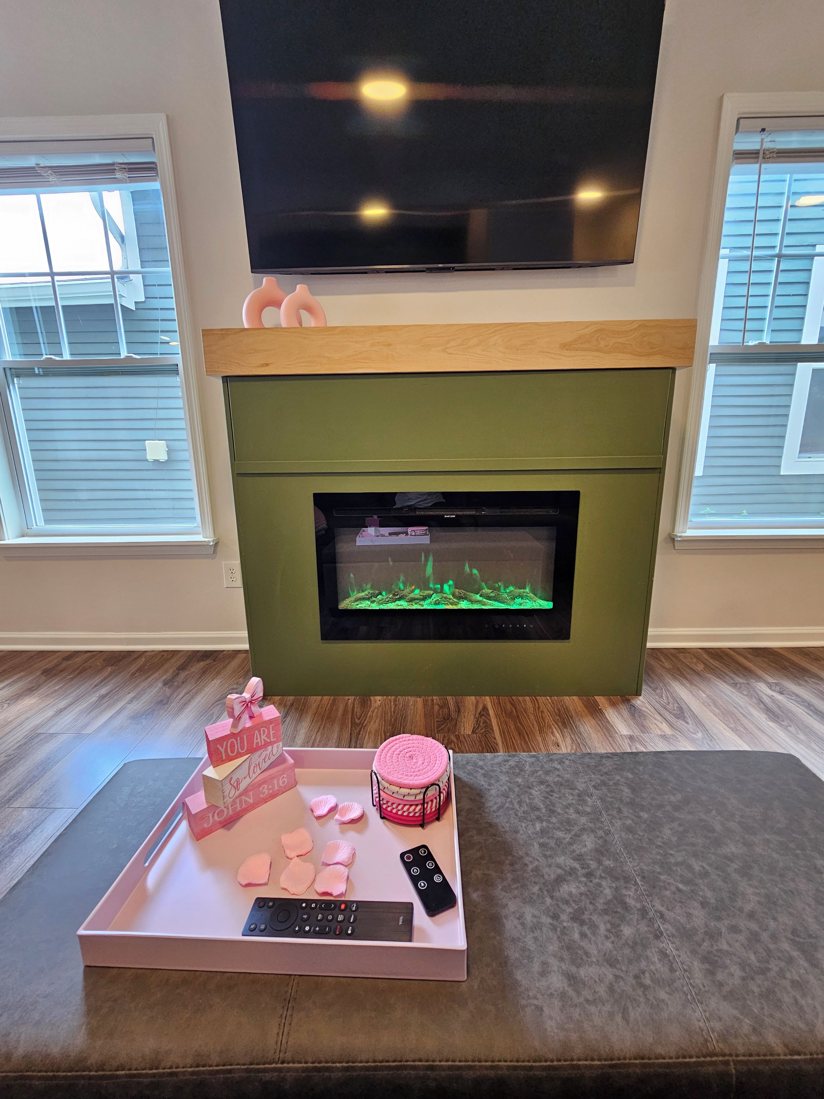 A living room with a flat-screen TV mounted above a green fireplace, a wooden mantel, and a tray with a pink box, seashells, and a remote control.