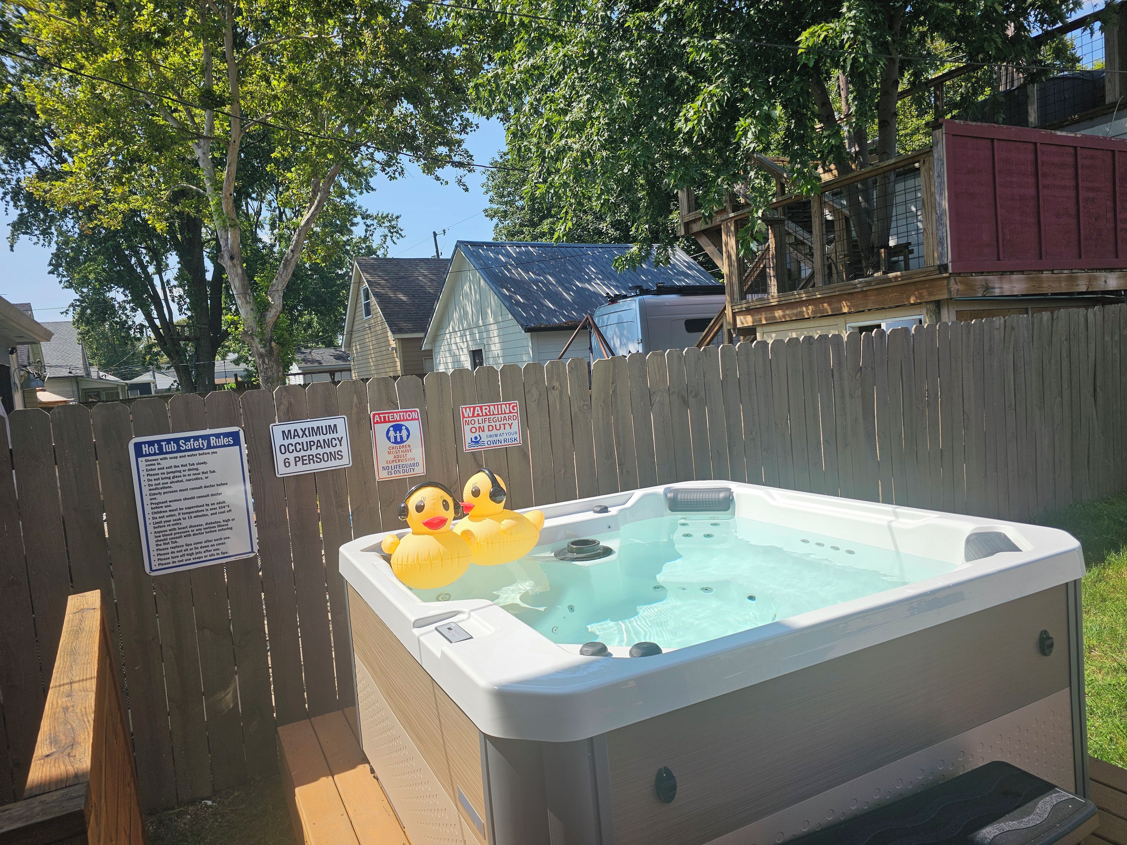 A hot tub with two rubber ducks, surrounded by a wooden fence, with safety signs and a house in the background.