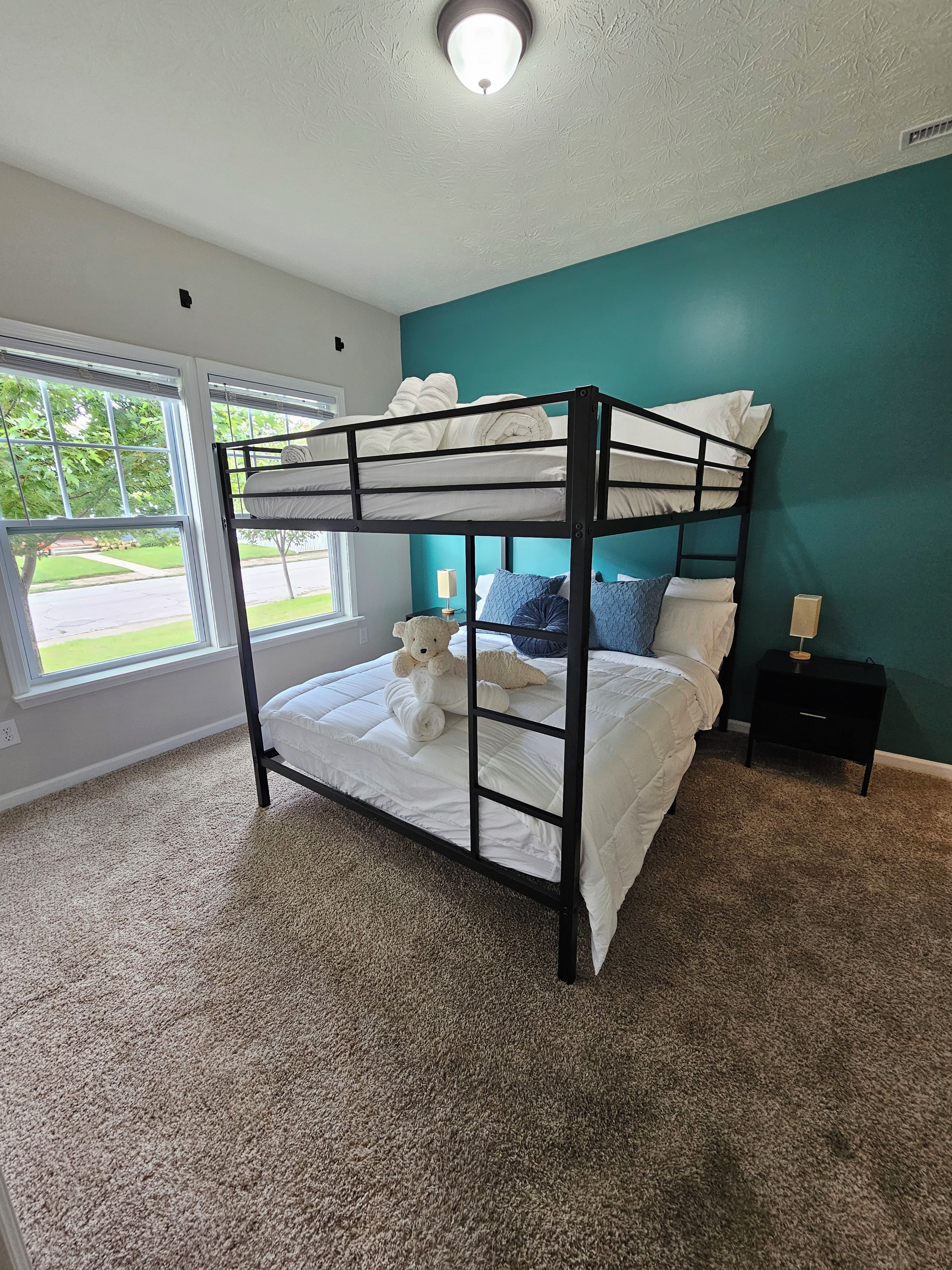 A bunk bed with a teddy bear, a nightstand, and a window with a view of greenery.