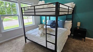 A bunk bed with a teddy bear, a nightstand, and a window with a view of greenery.