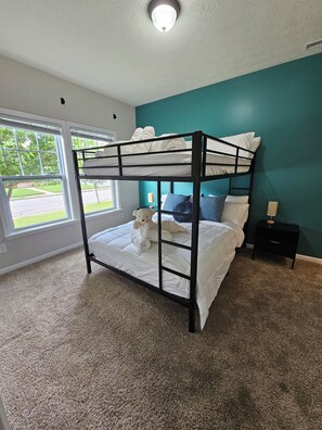Uma cama beliche com um urso de pelúcia, uma mesa de cabeceira e uma janela com vista para o verde.