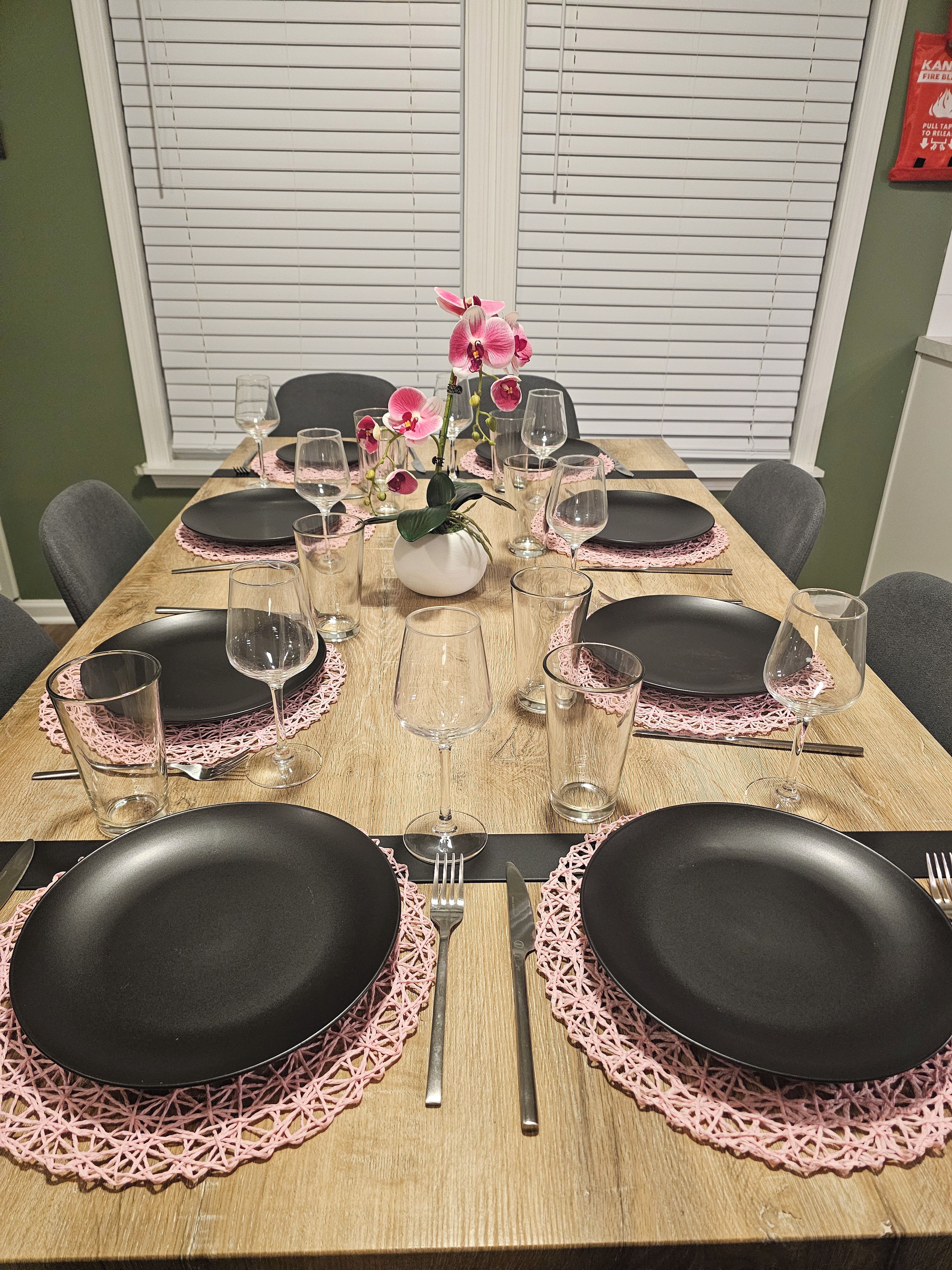 A dining table set for six with black plates, pink doilies, and clear wine glasses.