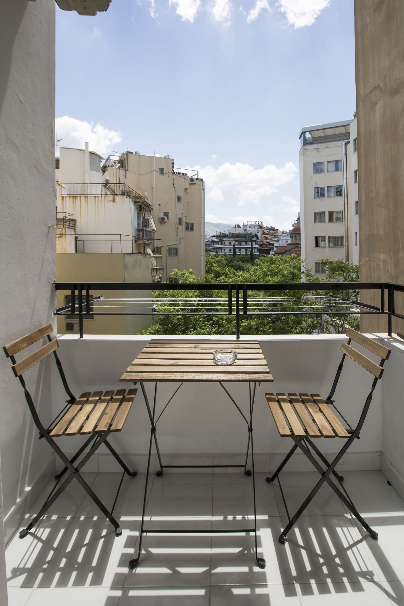 A balcony with a wooden table and two chairs, overlooking a cityscape with buildings and greenery.
