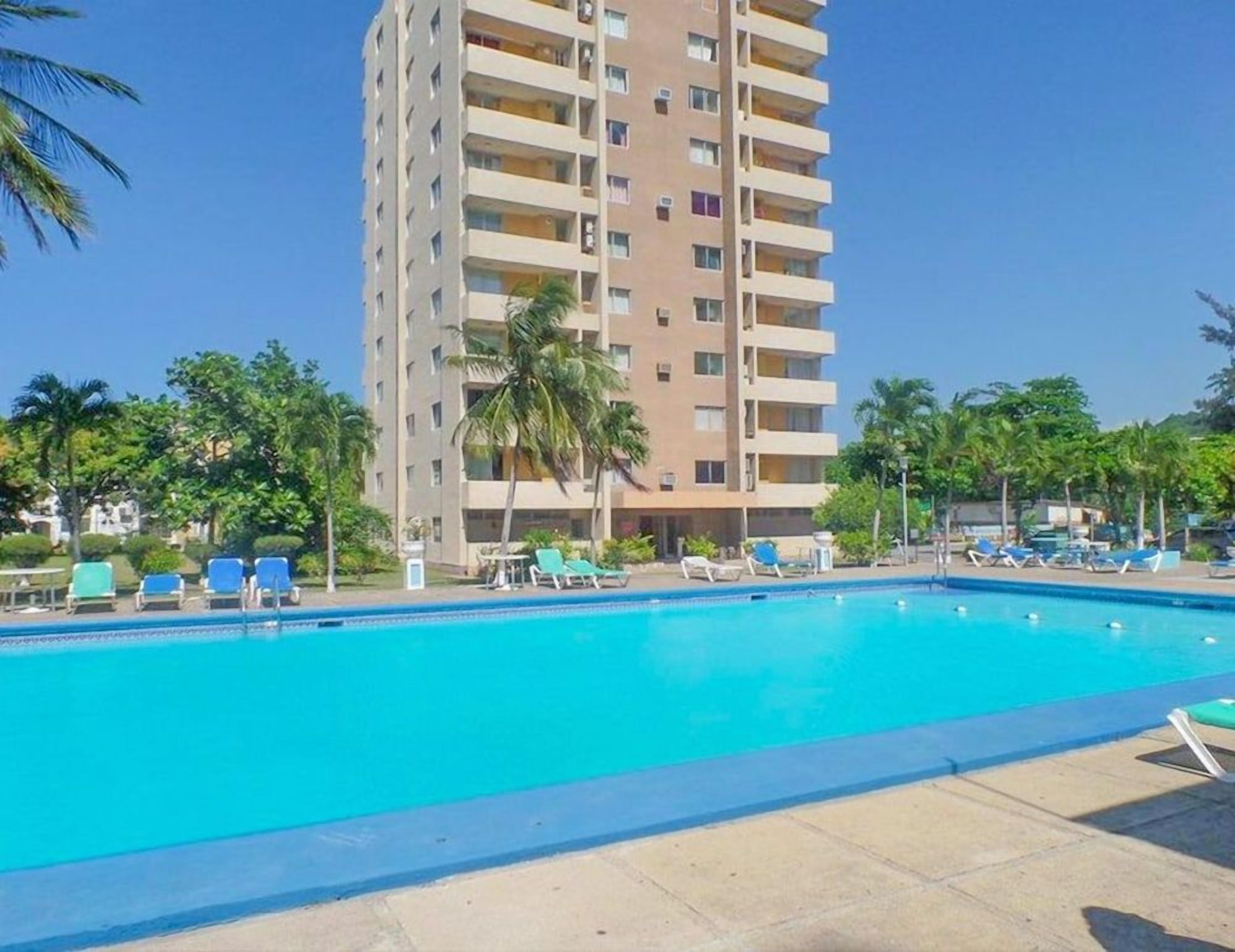Piscina com espreguiçadeiras, um edifício alto ao fundo e palmeiras.