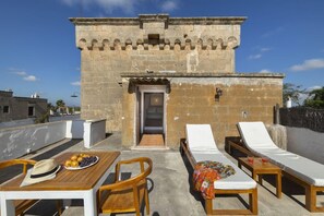 A rooftop terrace with a stone building, a table with a hat and fruit, and two sun loungers.
