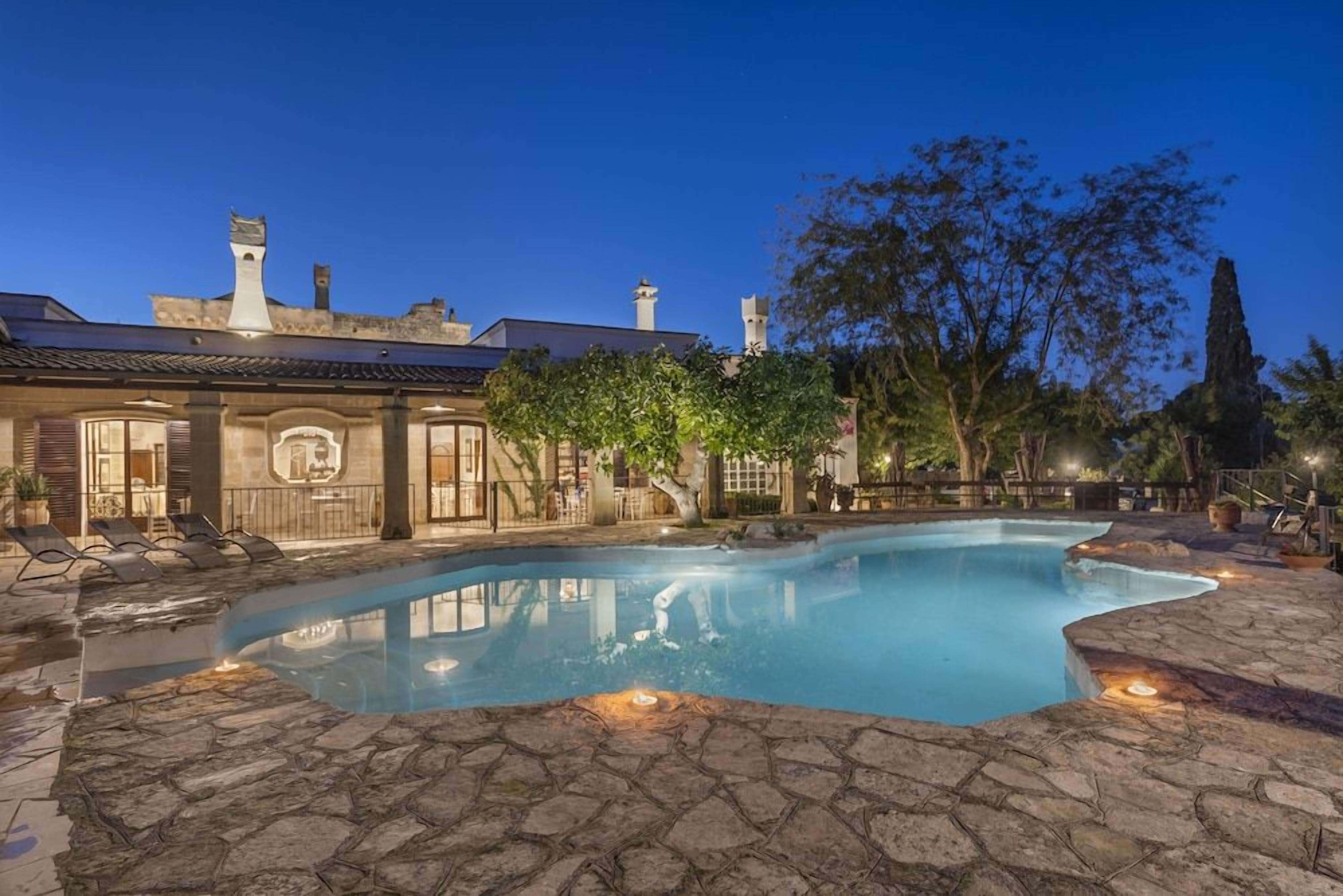 A swimming pool with stone tiles, a lounge area with chairs, and a building with a tiled roof and chimneys.