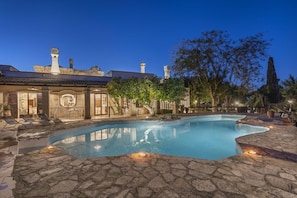 A swimming pool with stone tiles, a lounge area with chairs, and a building with a tiled roof and chimneys.