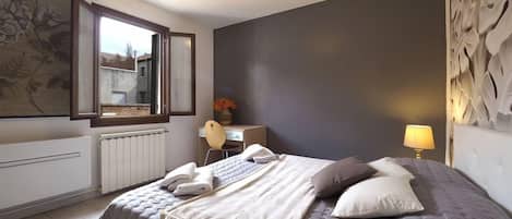 A bedroom with a large bed, a desk, a chair, a radiator, and a window with a view.