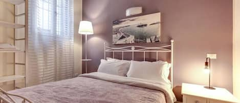 A bedroom with a bed, a bedside table, a lamp, a wall-mounted air conditioner, and a window with sheer curtains.