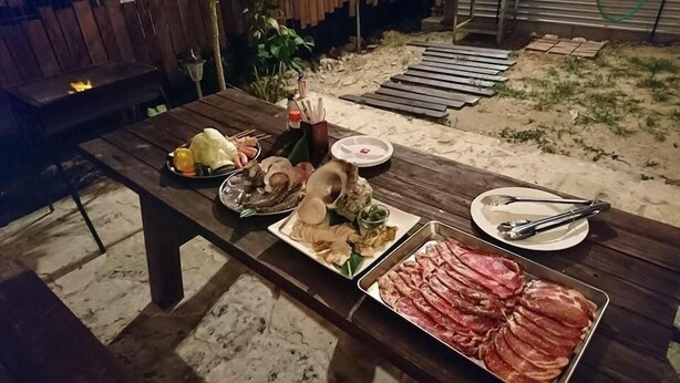 Una mesa de madera al aire libre con diversos alimentos, como embutidos y verduras, y una parrilla al fondo.