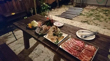 Una mesa de madera al aire libre con diversos alimentos, como embutidos y verduras, y una parrilla al fondo.