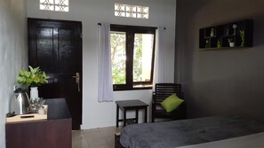 A room with a bed, a small table, a chair, a desk with a kettle, and a window with curtains. - Indah Homestay (Senggigi)