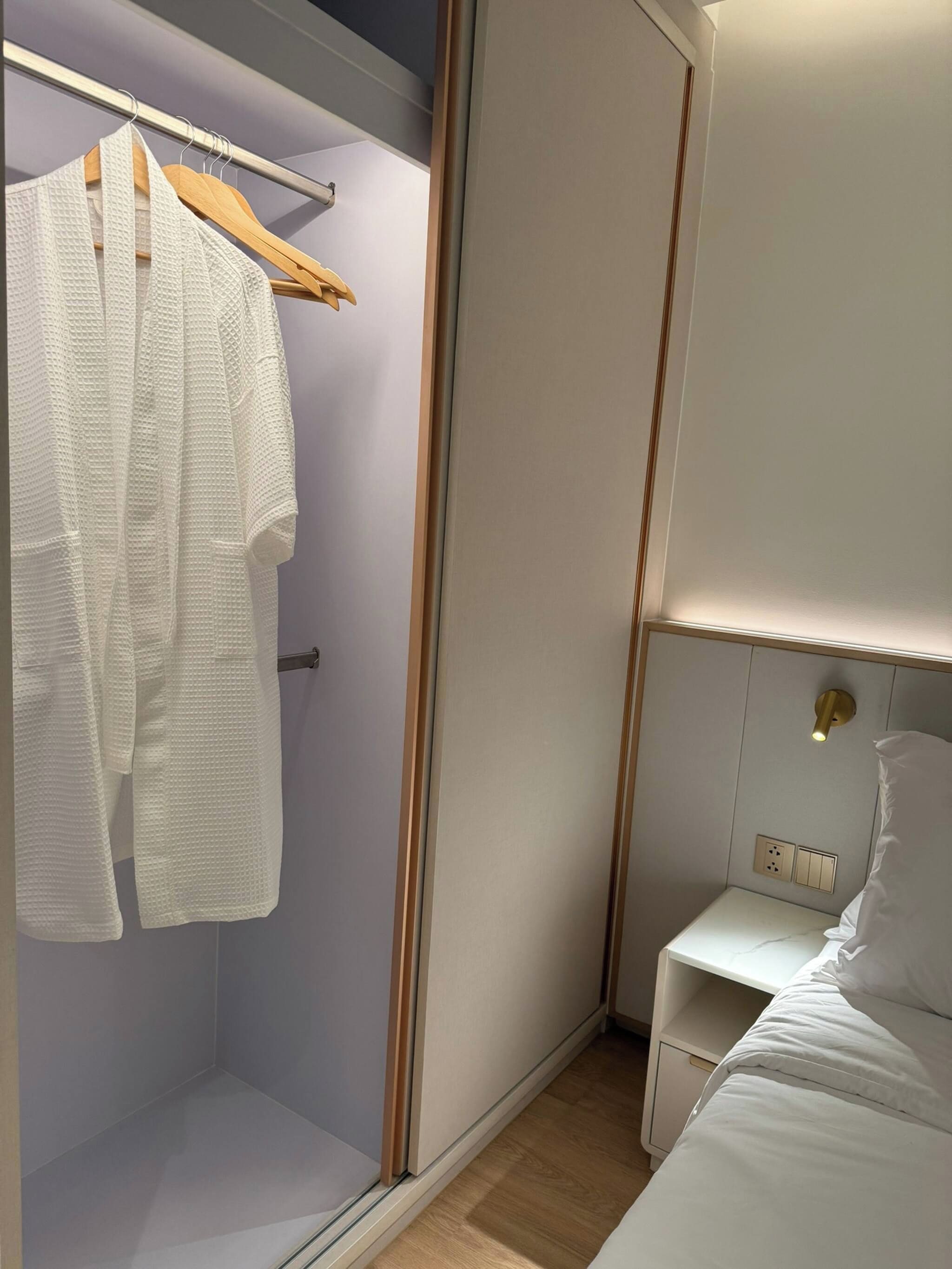 Een hotelkamer met een inloopkast, een bed met wit beddengoed, een nachtkastje en een wandlamp.