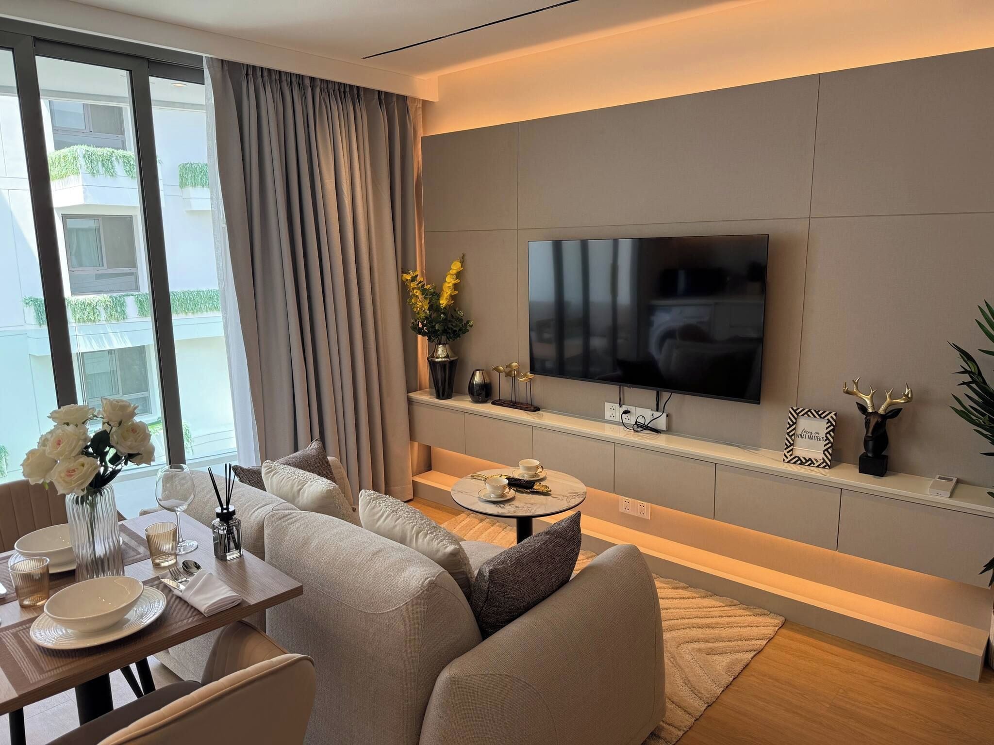 A modern living room with a sofa set, a coffee table, a flat-screen TV mounted on the wall, and a dining area with a table set for two.
