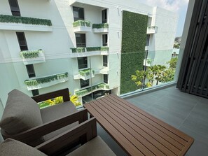 Un espacio moderno para sentarse al aire libre con una mesa y sillas de madera, barandilla de cristal y vistas a un edificio con jardines verticales verdes.