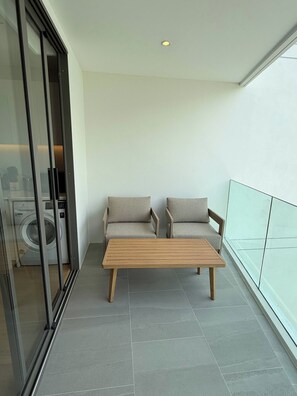 Ein Balkon mit zwei gepolsterten Stühlen und einem Holztisch, im Hintergrund eine Waschmaschine und ein Glasgeländer.
