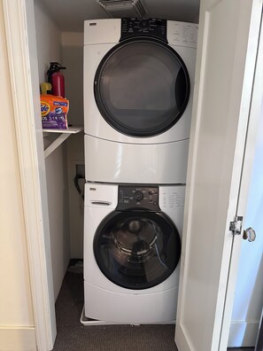 A compact laundry room with two stacked front-loading washing machines and a shelf with laundry detergent. - Charming Queen Anne Craftsman - Walk to Space Needle (Seattle)