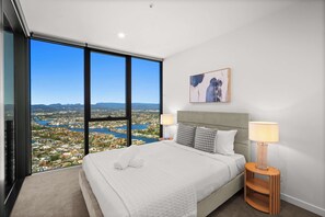 Ein Hotelzimmer mit einem großen Bett, zwei Nachttischlampen, Blick auf eine Stadtkulisse durch raumhohe Fenster und einem Gemälde an der Wand.