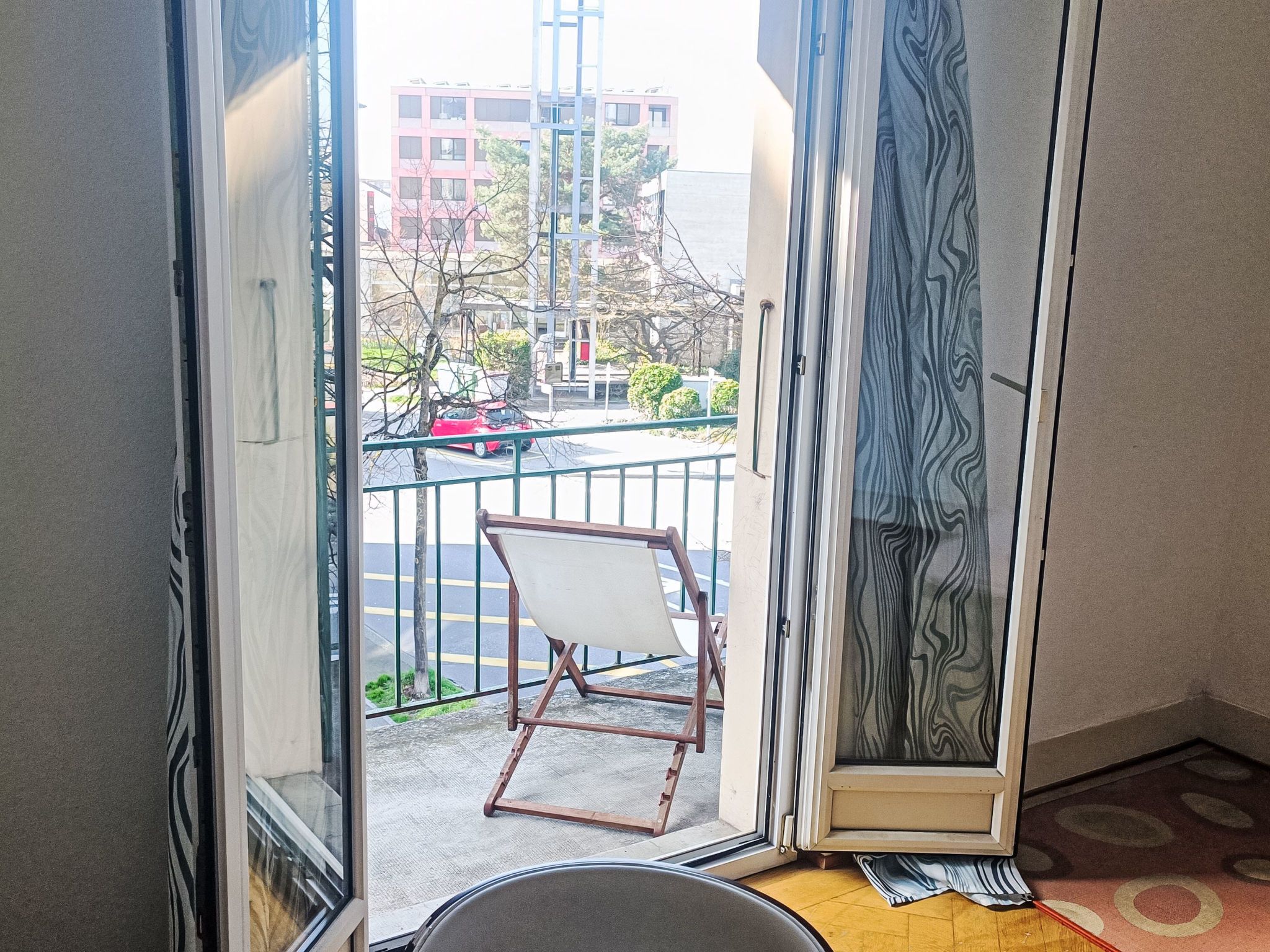 Ein Zimmer mit Blick auf eine Straße, ein Stuhl und ein gemustertes Vorhang.
