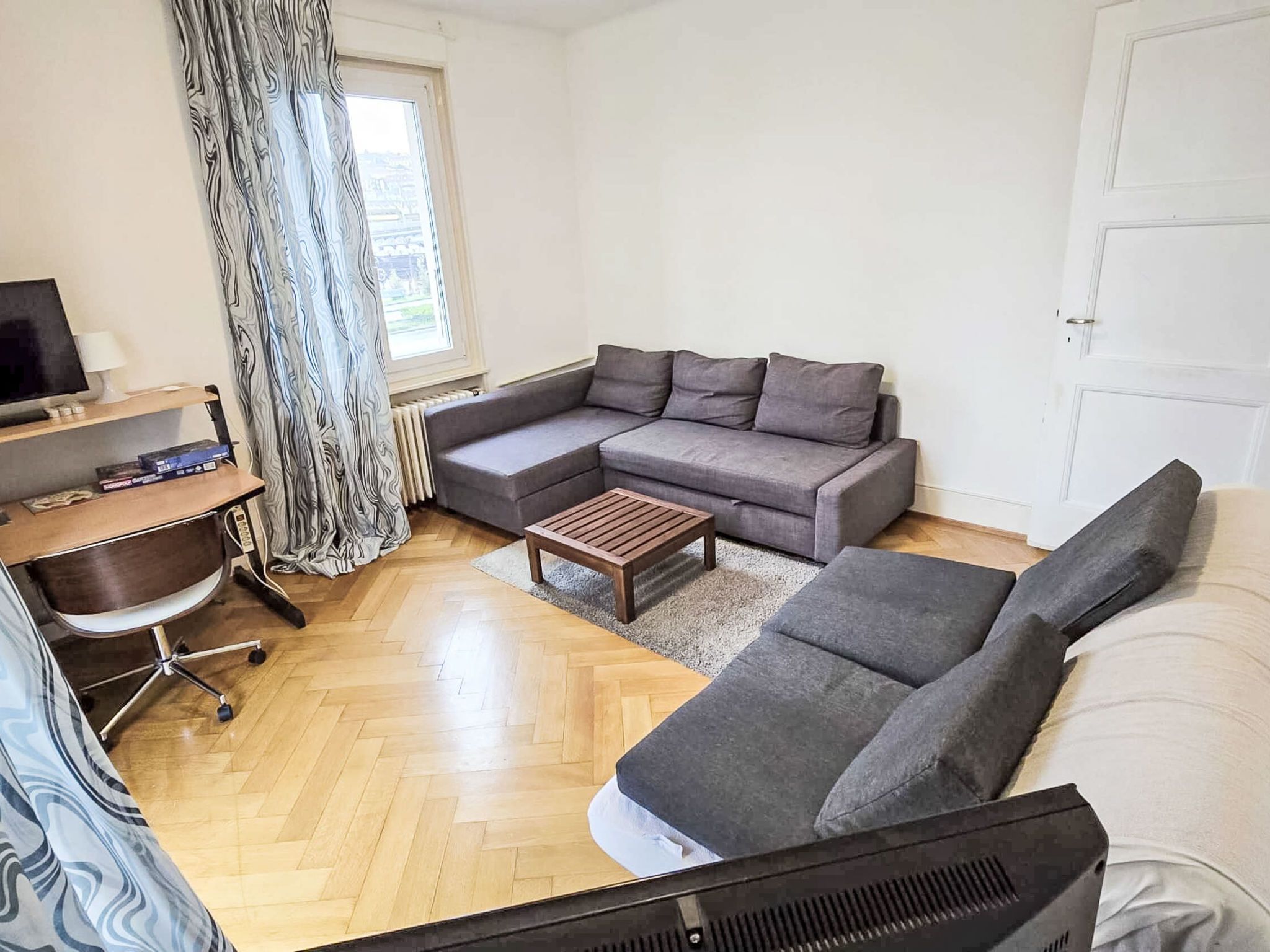 Ein modernes Wohnzimmer mit einem Ecksofa, einem Holztisch, einem Fernseher und einem Schreibtisch mit Stuhl.