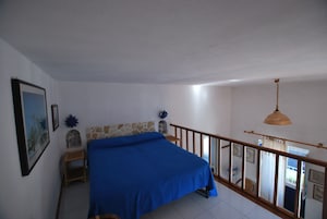 Un dormitorio con cama, mesita de noche de madera, pared de acento de piedra, balcón con barandilla y lámpara colgante.