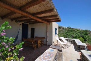 Um alpendre coberto com mesa e bancos de madeira, parede de pedra com padrão em azulejos e vista para uma encosta.