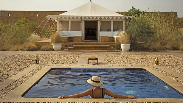 Piscina com uma pessoa sentada na borda, área de estar coberta com degraus que levam a uma porta e um edifício ao fundo.