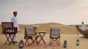 Cena de deserto com um camelo, um homem e uma mesa com cadeiras e lanternas.
