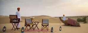Une scène de désert avec un chameau, un homme, une table, des chaises et des lanternes.