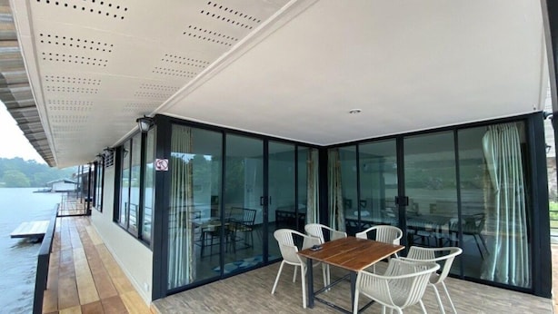 An outdoor dining area with a wooden table and white chairs, large glass windows, and a wooden deck. - Goodview Resort & Camping (Sai Yok)