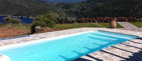 Una piscina rectangular con agua azul cristalina, rodeada de baldosas de piedra, con montañas y una masa de agua al fondo.