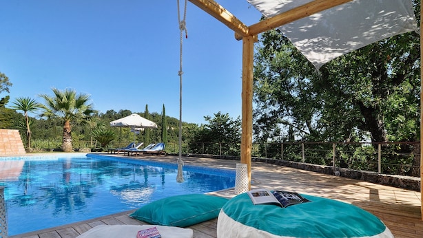Un área junto a la piscina con sillones de descanso, una terraza de madera y una pérgola con toldo blanco.