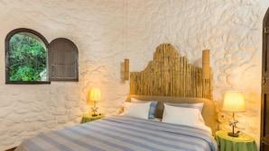 A bedroom with a stone wall, a bed with striped bedding, two bedside tables with lamps, and a window with shutters. - Villa Bournella (Corfu)