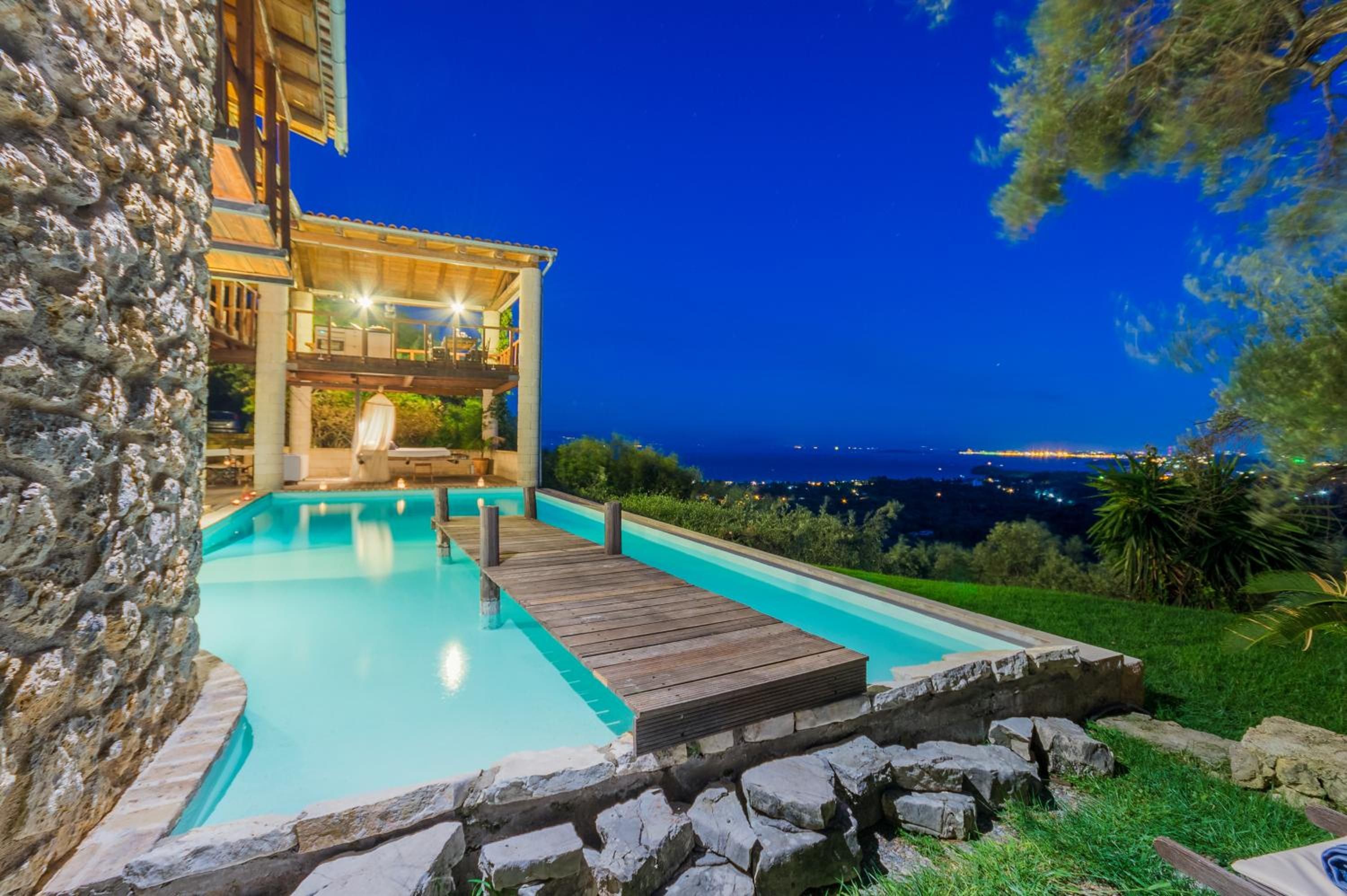 A pool with a wooden deck, a stone wall, and a view of the city at night.