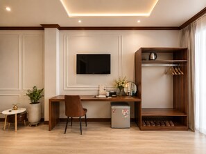 A modern hotel room with a wooden desk, a flat-screen TV mounted on the wall, a small refrigerator, a shoe rack, and a potted plant. - KAS Ancient Boutique Hotel (Ben Thanh Ward)