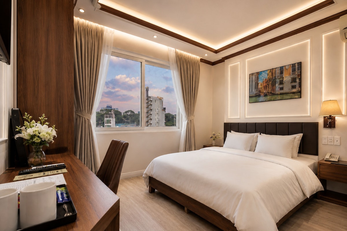 Ein Hotelzimmer mit einem Bett, einem Schreibtisch mit Blumen in einer Vase, einem Gemälde an der Wand und Blick auf Gebäude vor dem Fenster.