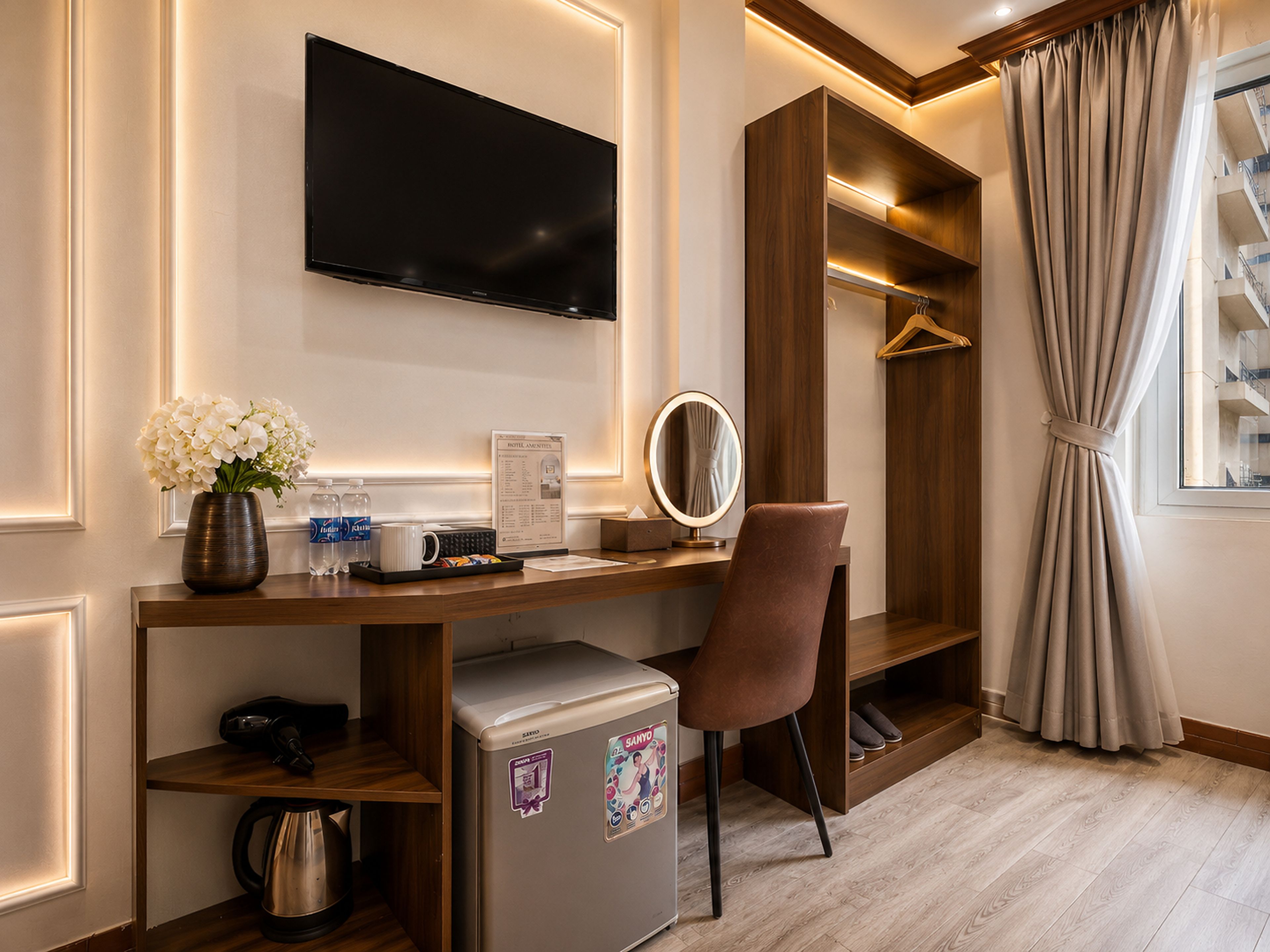 A hotel room with a flat-screen TV, a vanity with a mirror, a minibar, a small refrigerator, and a wooden desk.