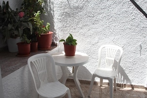 Un pequeño espacio al aire libre con dos sillas de plástico blancas y una mesa, rodeado de plantas en macetas.