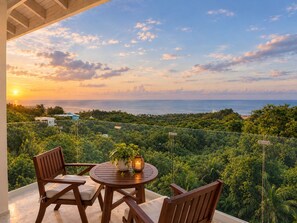 Ein Holztisch und Stühle auf einem Balkon mit Blick auf eine üppige Landschaft und den Sonnenuntergang über dem Meer.