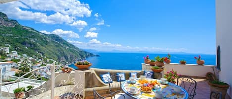 A terrace with a table set for breakfast, overlooking a coastal landscape with buildings and a clear sky.