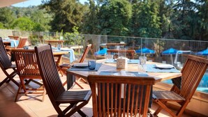 An outdoor dining area with wooden tables and chairs, a glass railing, and a view of trees and a pool. - Trypiti Hotel and Suites (Thasos)