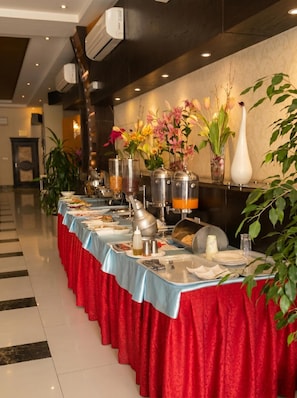 A buffet setup with a long table covered in a red tablecloth, various food containers, and decorative vases with flowers. - Tulip Al Rawdah (Riyadh)