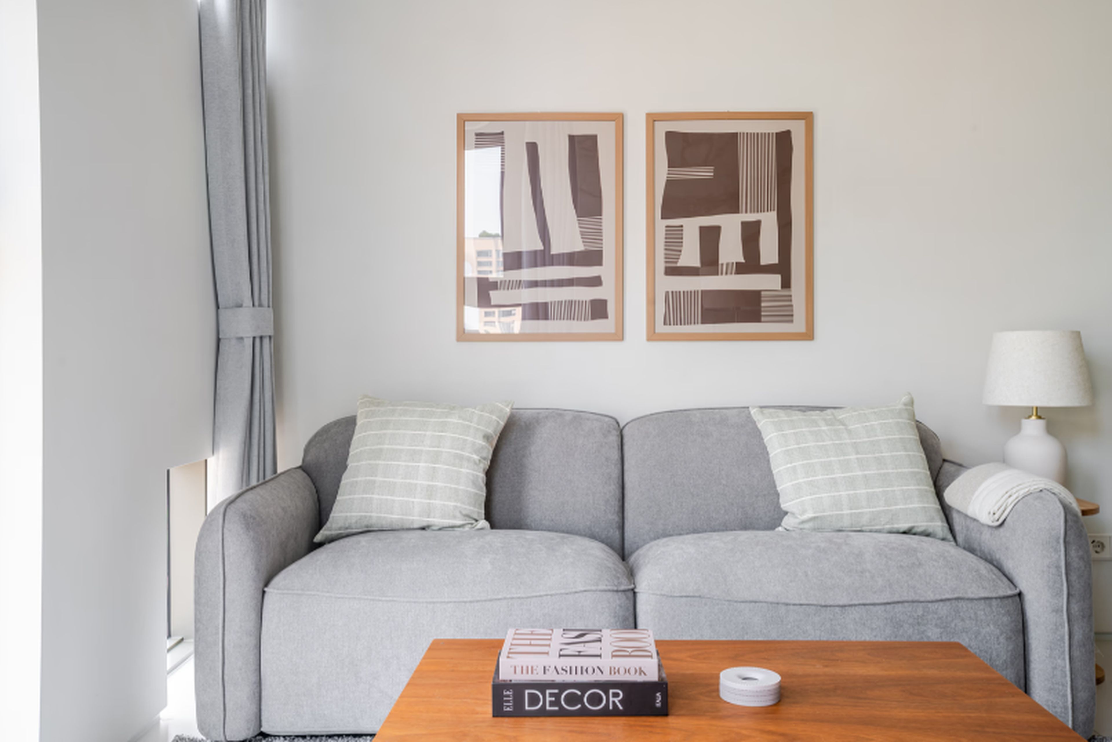 Uma sala de estar moderna com um sofá cinzento, duas almofadas, uma mesa de centro de madeira com livros e duas obras de arte abstrata emolduradas na parede.