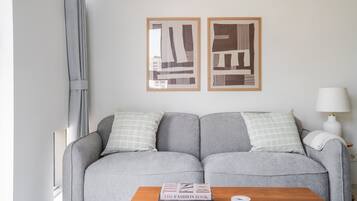 Uma sala de estar moderna com um sofá cinzento, duas almofadas, uma mesa de centro de madeira com livros e duas obras de arte abstrata emolduradas na parede.