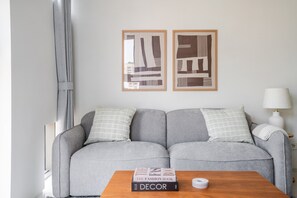 Uma sala de estar moderna com um sofá cinzento, duas almofadas, uma mesa de centro de madeira com livros e duas obras de arte abstrata emolduradas na parede.
