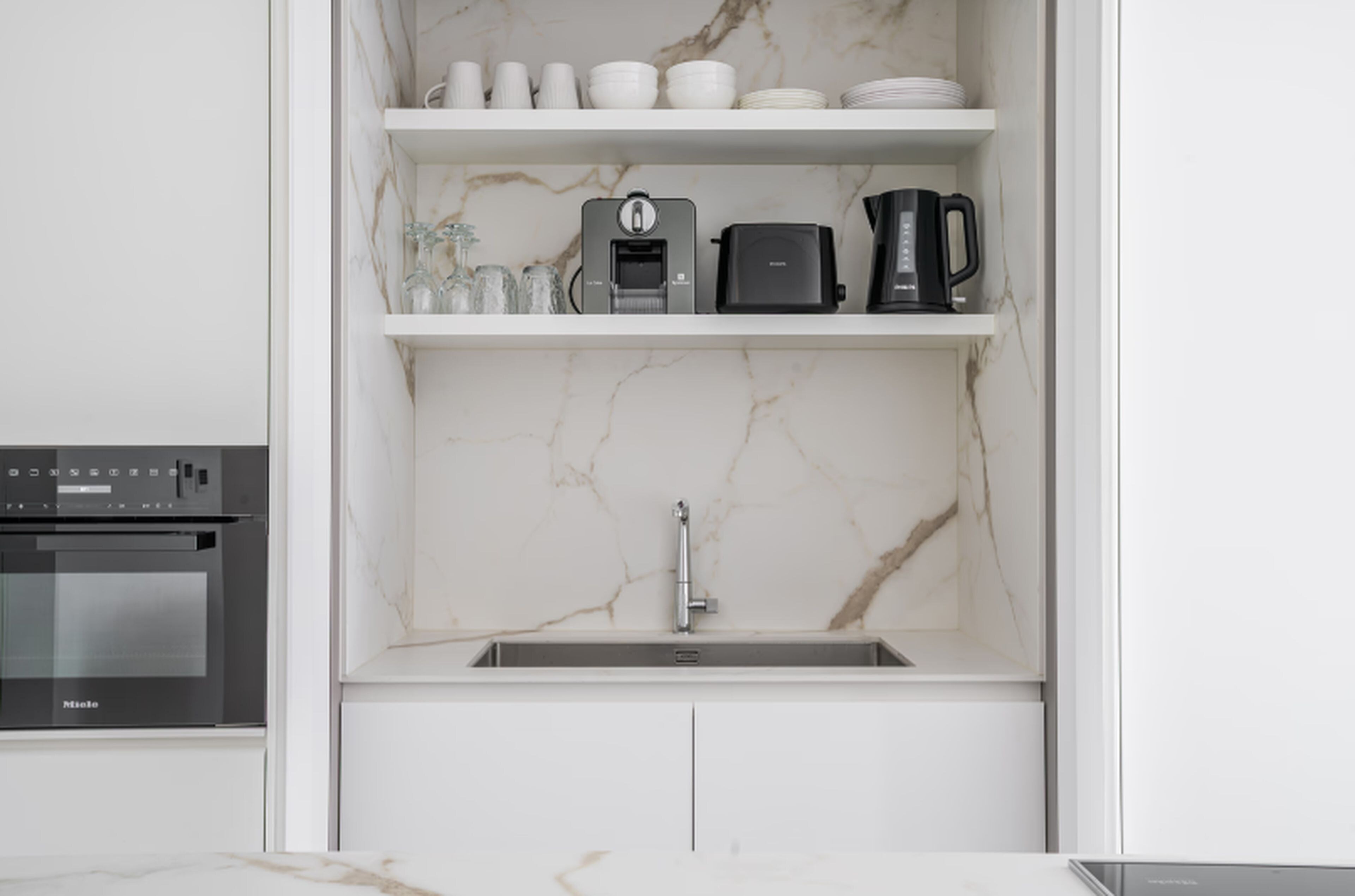 A kitchen with a marble backsplash, a built-in oven, a sink, and a shelf with kitchenware and appliances.