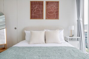 A hotel room with a bed, two wall art pieces, a nightstand with a lamp, and a view of the outside.